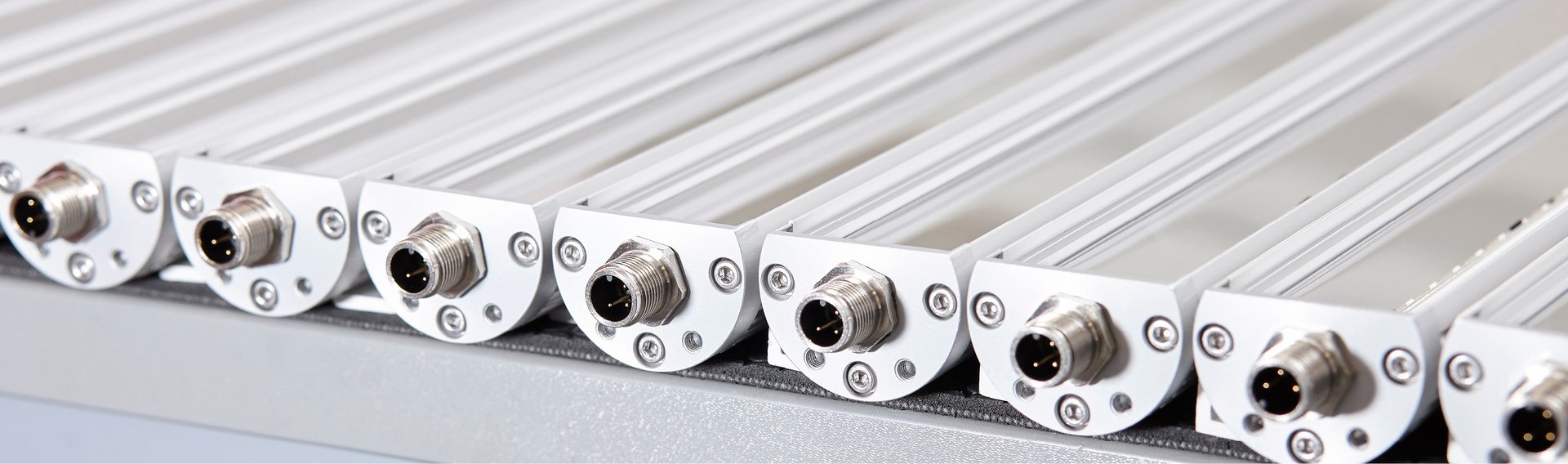 Serie de luminarias LED industriales montadas muy juntas, cada una con un conector metálico M12 en la parte frontal, fijadas sobre perfiles de aluminio claros.