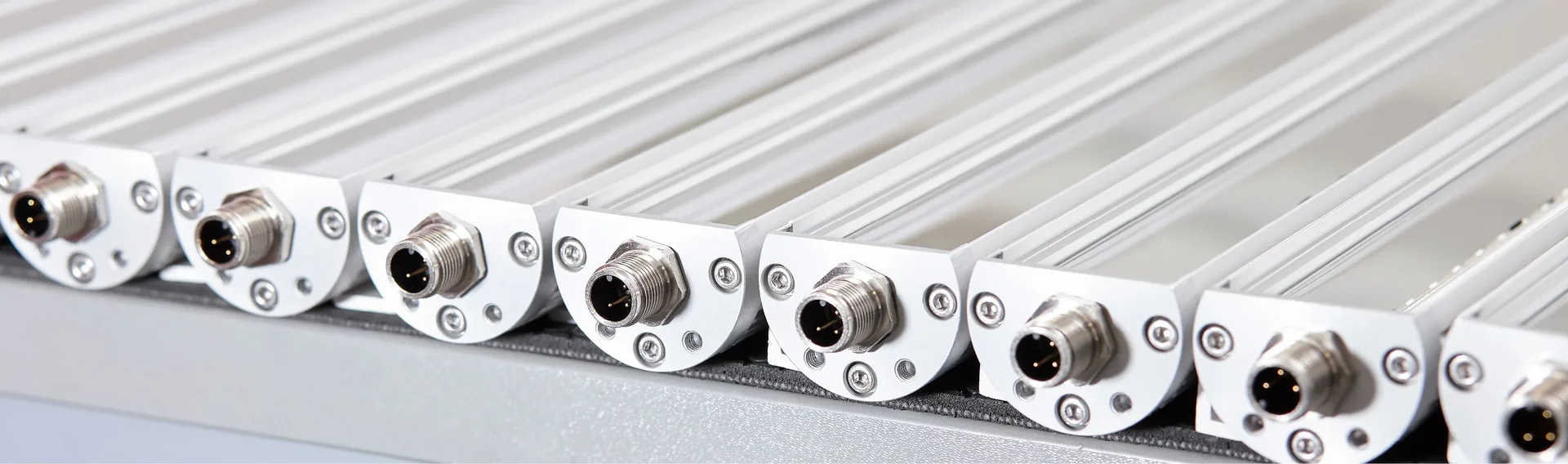 Series of industrial LED fixtures mounted closely side by side, each equipped with a metallic M12 connector on the front, installed on bright aluminum profiles.
